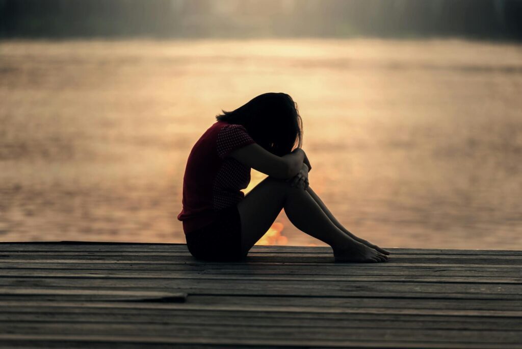 Mujer sentada sola en un muelle al atardecer con la cabeza apoyada en sus rodillas, simbolizando la tristeza y la introspección.