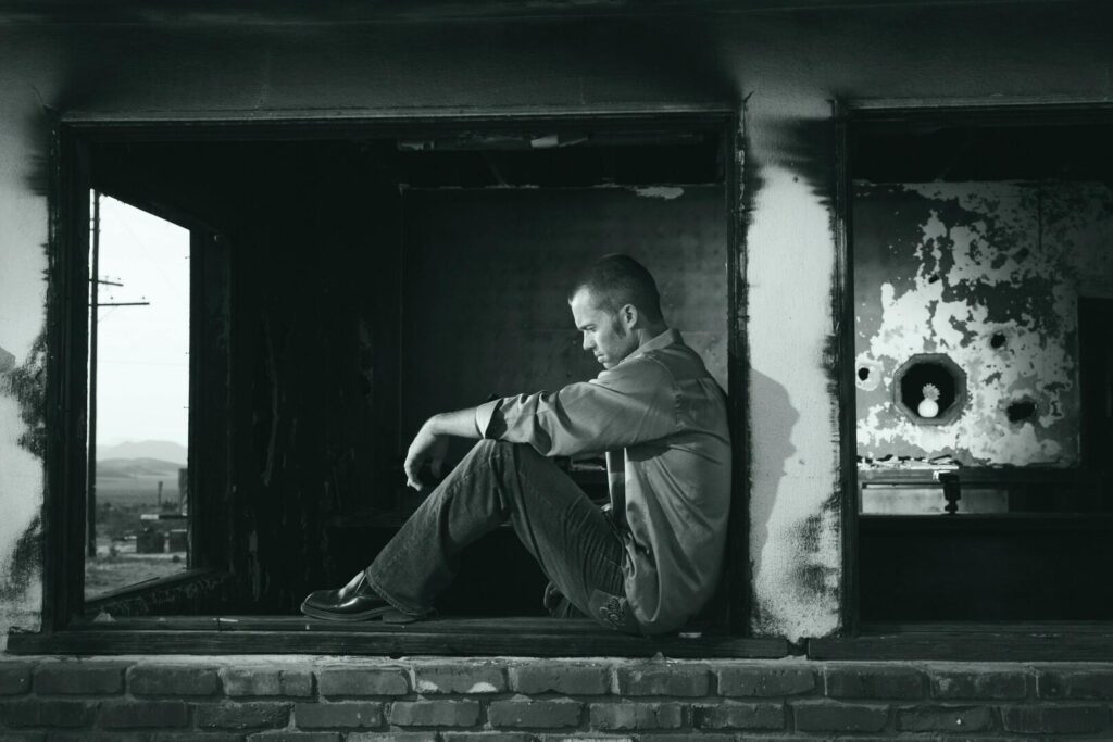 Hombre sentado solo en el alféizar de una ventana en un edificio en ruinas, con expresión de tristeza y reflexión.