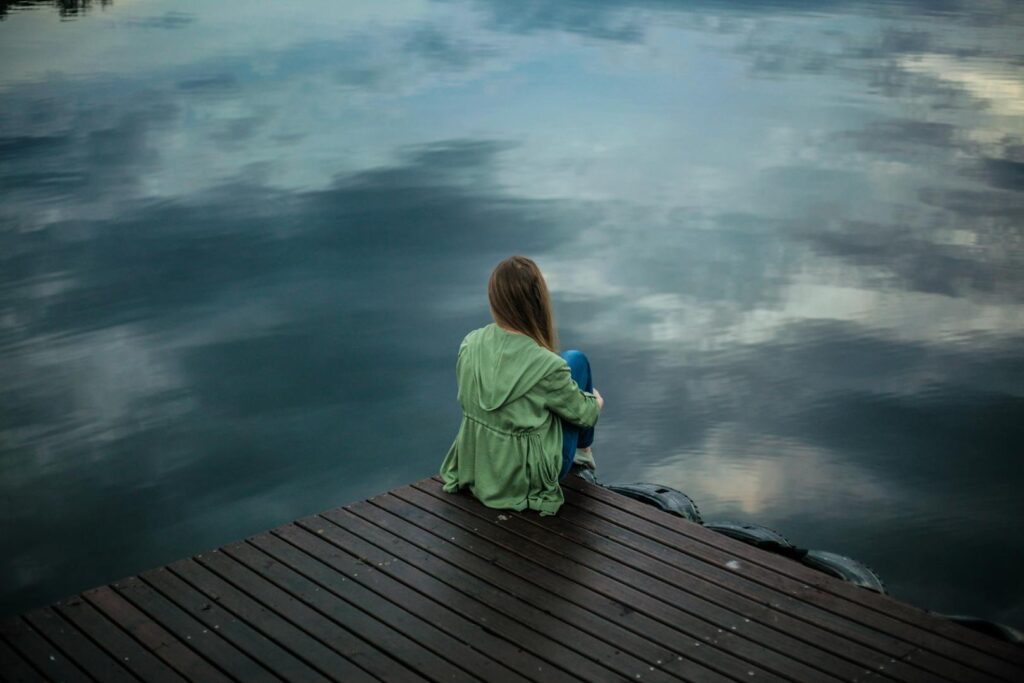 Mujer sentada sola en un muelle mirando hacia el agua en un día nublado, simbolizando la reflexión y la tranquilidad.